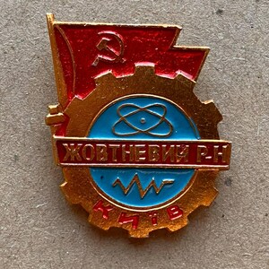 May include: A gold-toned metal pin featuring a red flag with a hammer and sickle, a gear, and a blue circular centre. The centre displays an atom symbol and a zigzag line. The text ЖОВТНЕВИЙ Р.Н and КИЇВ is printed in red on the metal surface.