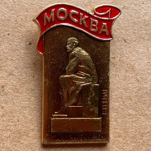 May include: A rectangular metal pin featuring a gold-toned relief of a seated figure on a pedestal. Above the figure, a red banner with gold trim displays the Cyrillic text "MOCKBA". The design includes additional small Cyrillic text at the base.