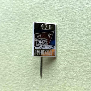 Op de afbeelding: Een rechthoekige speld met een zwarte en blauwe achtergrond en het jaartal 1970 bovenaan. Het ontwerp bevat een witte illustratie van de Lunokhod 1 maanrover op een oppervlak met kraters, met de cyrillische tekst LUNOKHOD 1 onderaan.