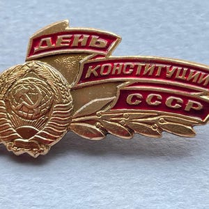 May include: A gold-colored Soviet-era pin with red accents. The pin features a circular emblem with a hammer and sickle, surrounded by wheat stalks. The text "День Конституции СССР" is displayed on red banners.