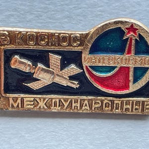 May include: Vintage gold-toned pin with Russian text and a space theme. The pin features a satellite, a star, and the words "B KOCMOC" and "MEЖДУHAPOДHЫE". The design includes red, blue, and green enamel.