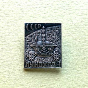 Puede incluir: Un pin rectangular de metal plateado con un diseño en relieve del vehículo lunar Lunokhod-1 sobre una superficie texturizada con cráteres. La esquina superior izquierda muestra el texto cirílico CCCP y el borde inferior el texto LUNOKHOD-1.