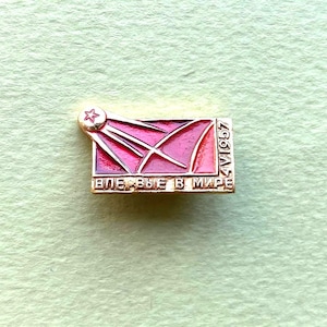 Puede incluir: Un broche rectangular de metal dorado con incrustaciones de esmalte rojo y un diseño estilizado de estrella fugaz. El broche incluye el texto en cirílico "ВПЕРВЫЕ В МИРЕ" en el borde inferior y la fecha "4.X.1957" verticalmente a la derecha.