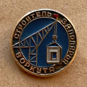 Puede incluir: Un pin metálico circular con fondo de esmalte azul que muestra una grúa de construcción dorada levantando un bloque de construcción blanco. El texto "СТРОИТЕЛЬ ЗАПОЛЯРЬЯ ВОРКУТА" está grabado en letras doradas alrededor del borde.