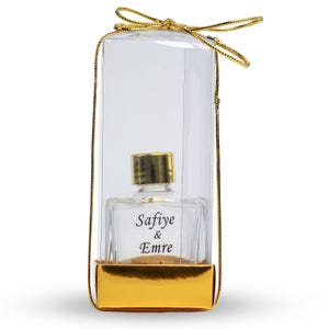 Op de afbeelding: Een heldere glazen parfumfles met een gouden dop, verpakt in een transparante plastic doos met een gouden basis. De fles heeft de namen "Safiye & Emre" erop gedrukt en is vastgebonden met een gouden lint.