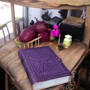 May include: A purple leather-bound journal with an embossed design featuring a figure, symbols, and a serpent. The journal is open on a wooden surface, with other decorative items in the background, including purple egg-shaped objects and a small green jar.