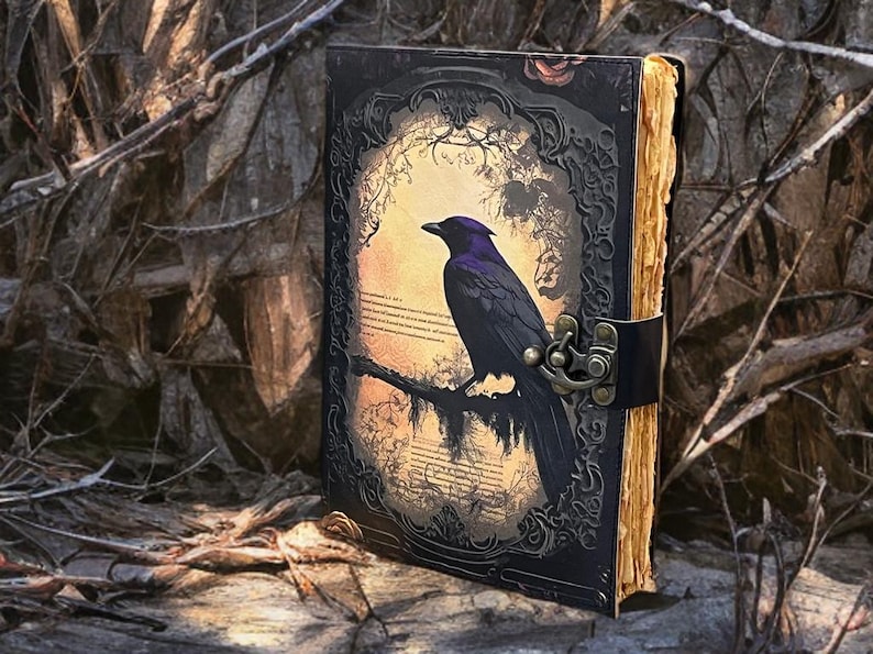 May include: A gothic journal with a black cover depicting a raven on a branch. The raven has purple detailing. The journal has a metal clasp and aged pages. The background is a blurred image of a forest, with a leather strap.
