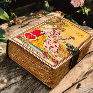 Puede incluir: Un diario de cartas del tarot de estilo vintage con arte de la carta "La Emperatriz". El diario tiene páginas envejecidas y texturizadas y un cierre decorativo. La portada representa a una mujer con un vestido fluido, un árbol frutal y el sol.