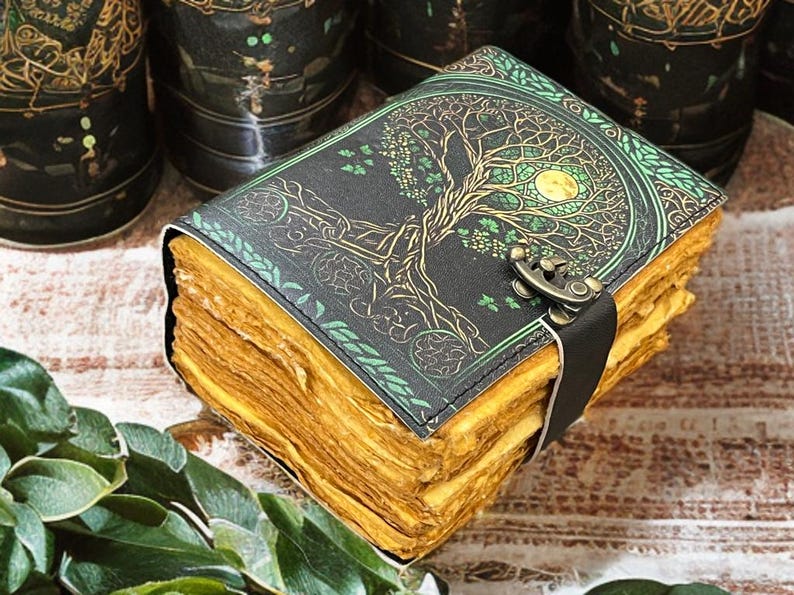 Puede incluir: Un diario encuadernado en cuero con un dise&ntilde;o de &aacute;rbol de la vida en relieve en la portada. La portada es negra con detalles dorados y verdes, y las p&aacute;ginas tienen un aspecto envejecido. Una correa negra con un cierre met&aacute;lico asegura el diario.