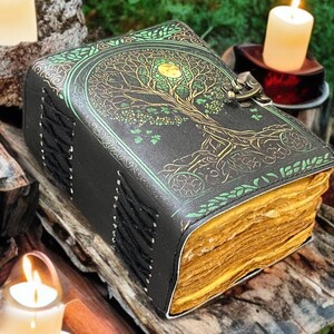 Puede incluir: Un diario de cuero negro con un dise&ntilde;o de &aacute;rbol de la vida en dorado y verde. El diario tiene un cierre de broche y encuadernaci&oacute;n cosida. Las p&aacute;ginas tienen un aspecto vintage y envejecido. El diario est&aacute; sobre un trozo de madera, con velas encendidas al fondo.