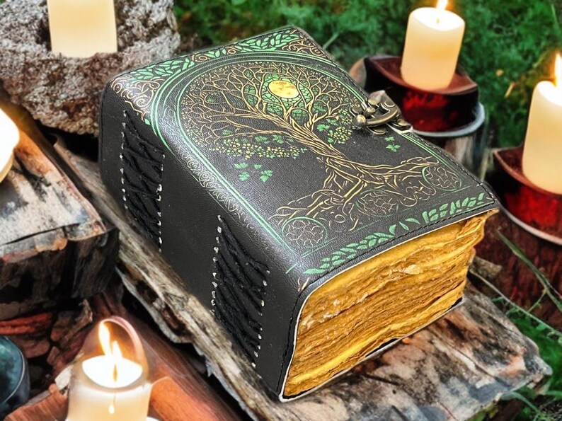 Puede incluir: Un diario de cuero negro con un dise&ntilde;o de &aacute;rbol de la vida en dorado y verde. El diario tiene un cierre de broche y encuadernaci&oacute;n cosida. Las p&aacute;ginas tienen un aspecto vintage y envejecido. El diario est&aacute; sobre un trozo de madera, con velas encendidas al fondo.
