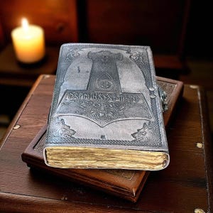 May include: A silver-colored leather journal with an embossed design featuring a hammer, runes, and wolf heads. The journal is closed with a clasp and rests on top of a brown leather-bound book. The pages have a vintage, aged appearance.