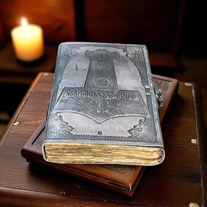 May include: A silver-colored leather journal with an embossed design featuring a hammer, runes, and wolf heads. The journal is closed with a clasp and rests on top of a brown leather-bound book. The pages have a vintage, aged appearance.