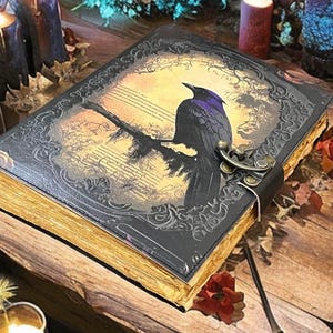 May include: A leather-bound journal with a raven illustration. The cover features a black raven perched on a branch against a yellowed background with musical notes. The edges of the pages are gold, and the journal is secured with a metal clasp.