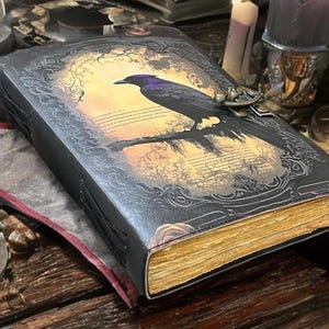 May include: A dark, embossed leather journal featuring a raven illustration on the cover. The raven sits on a branch against a sunset backdrop. The pages have a golden hue, and the journal is secured with a metal clasp. The overall theme is gothic and fantasy.