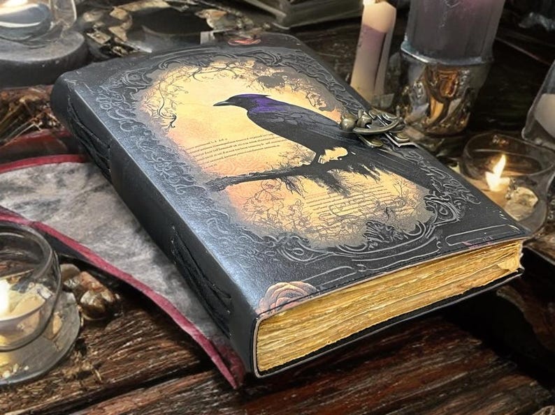 May include: A dark, embossed leather journal featuring a raven illustration on the cover. The raven sits on a branch against a sunset backdrop. The pages have a golden hue, and the journal is secured with a metal clasp. The overall theme is gothic and fantasy.