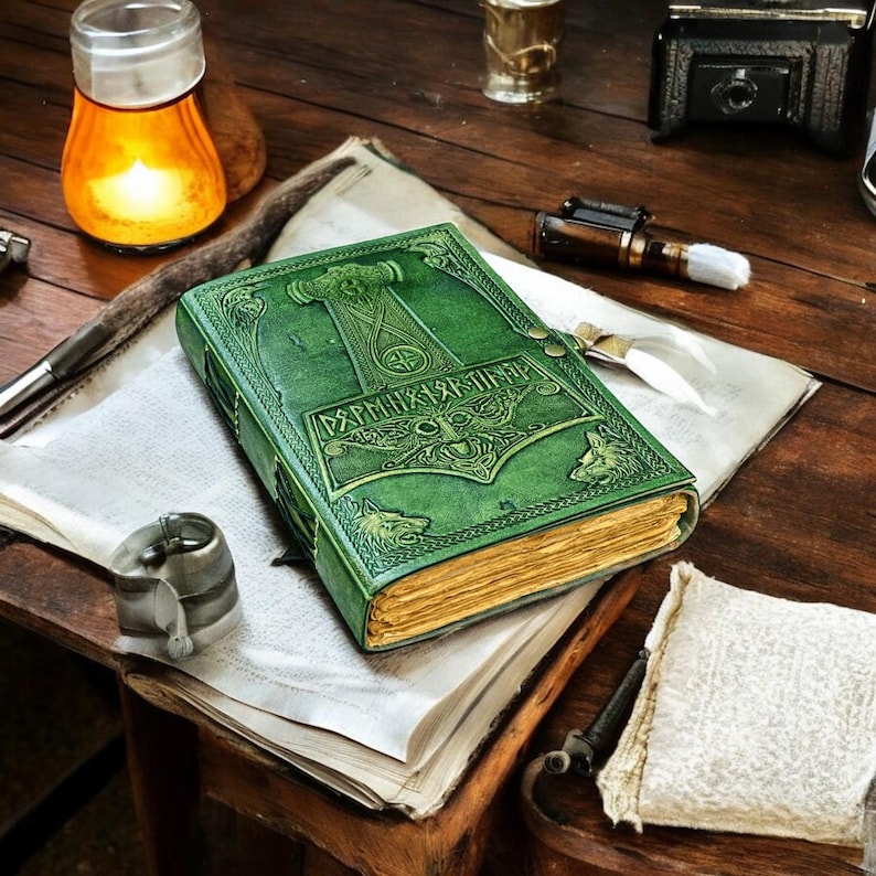 May include: A green leather-bound journal with embossed Norse designs, including a hammer and wolf figures. The journal has gold-edged pages and a clasp closure. The cover features runic text. The journal rests on a vintage wooden surface.