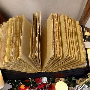 Puede incluir: Un libro abierto y envejecido con p&aacute;ginas gruesas y amarillentas. El libro est&aacute; rodeado de velas encendidas y elementos decorativos. Las p&aacute;ginas tienen un aspecto rugoso y texturizado, lo que sugiere edad y uso. La est&eacute;tica general es vintage y m&iacute;stica.