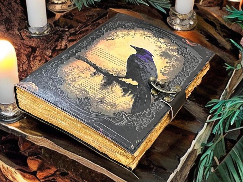 May include: A leather-bound journal featuring a raven illustration. The black cover has an embossed design and a metal clasp. The pages have a golden edge. The raven sits on a branch against a sepia-toned background.
