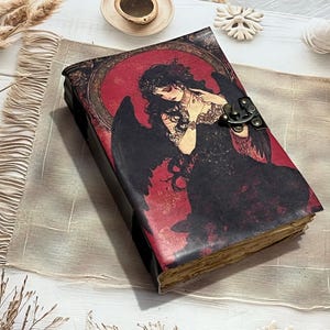 May include: A rectangular journal with a dark cover featuring a central illustration of a winged figure in a black dress against a red circular background. The book includes a metallic bronze-toned latch closure and visible aged, textured paper pages.