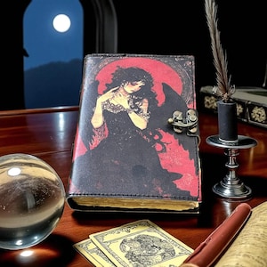 May include: A gothic journal with a black and red cover depicting a woman with black wings. The journal has a metal clasp and aged pages. A crystal ball, cards, and a quill pen are also visible, creating a mystical scene.