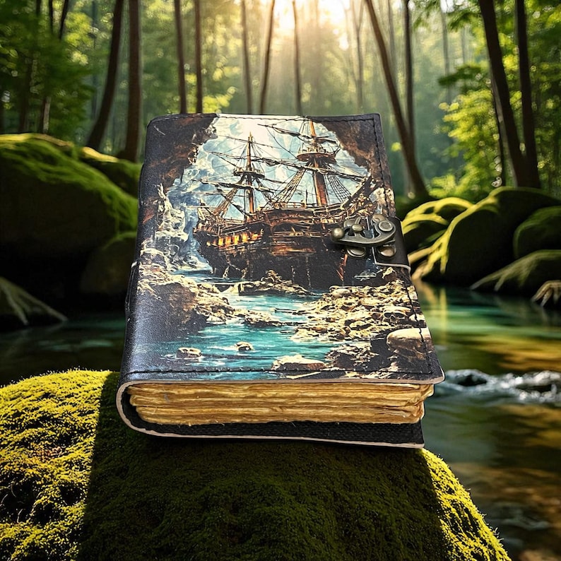 May include: A leather-bound journal with a detailed illustration of a tall ship sailing through a rocky cove. The cover features a metal clasp. The pages have a golden hue, and the journal is set against a mossy background.