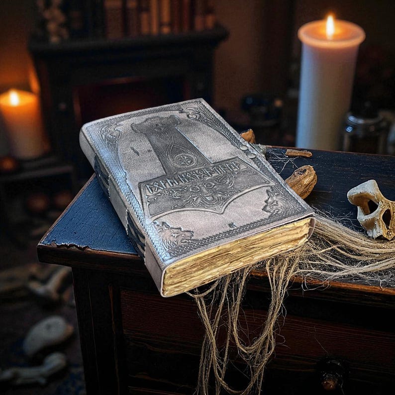 May include: A silver-colored leather-bound journal with an embossed design featuring a hammer and runes. The journal has aged, yellowed pages and is resting on a dark wooden surface. The cover also features wolf designs.