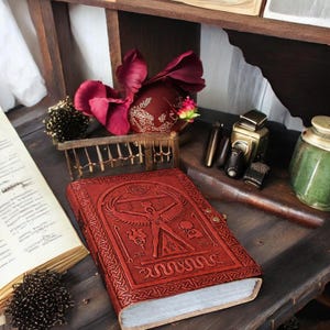 May include: A red leather-bound journal with an embossed design featuring a human figure and serpent. The journal is on a wooden surface with other items, including a pen, inkwell, and decorative flowers. The journal's cover has intricate patterns.