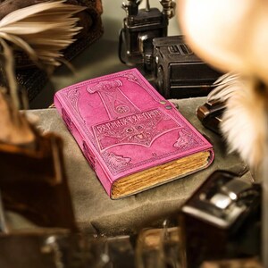 May include: A pink leather journal with embossed Norse mythology designs. The cover features a hammer, runes, and wolf heads. The journal has aged, yellowed pages and a metal clasp closure. The overall theme is fantasy and historical.