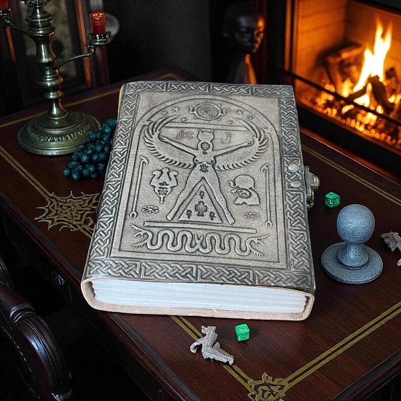 May include: A large, embossed leather-bound book with intricate designs, including a central figure with outstretched arms and wings. The cover is a light brown color with a metal clasp. The book is on a wooden table with other decorative items.