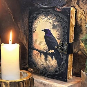 May include: A leather-bound journal with a raven illustration. The black cover has an ornate frame and a metal clasp. Lit candles in holders and a wooden base are in the foreground, creating a mystical ambiance.