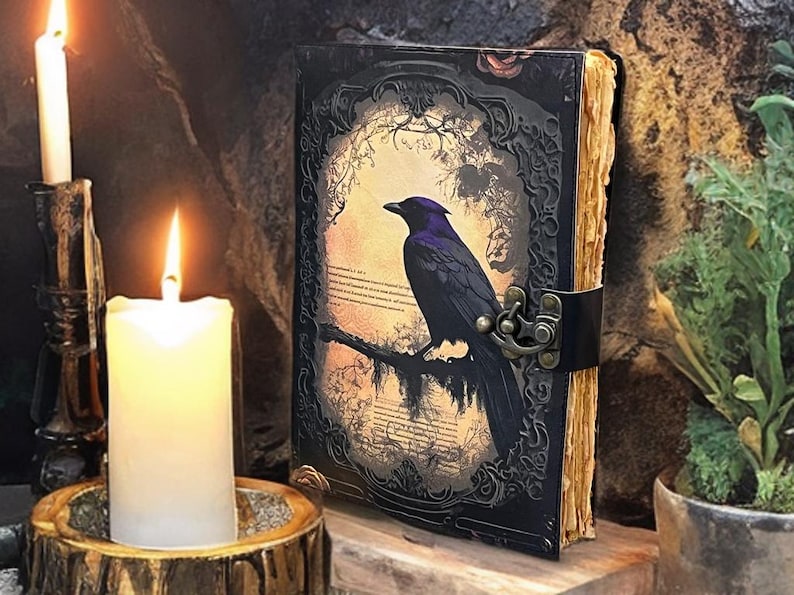 May include: A leather-bound journal with a raven illustration. The black cover has an ornate frame and a metal clasp. Lit candles in holders and a wooden base are in the foreground, creating a mystical ambiance.