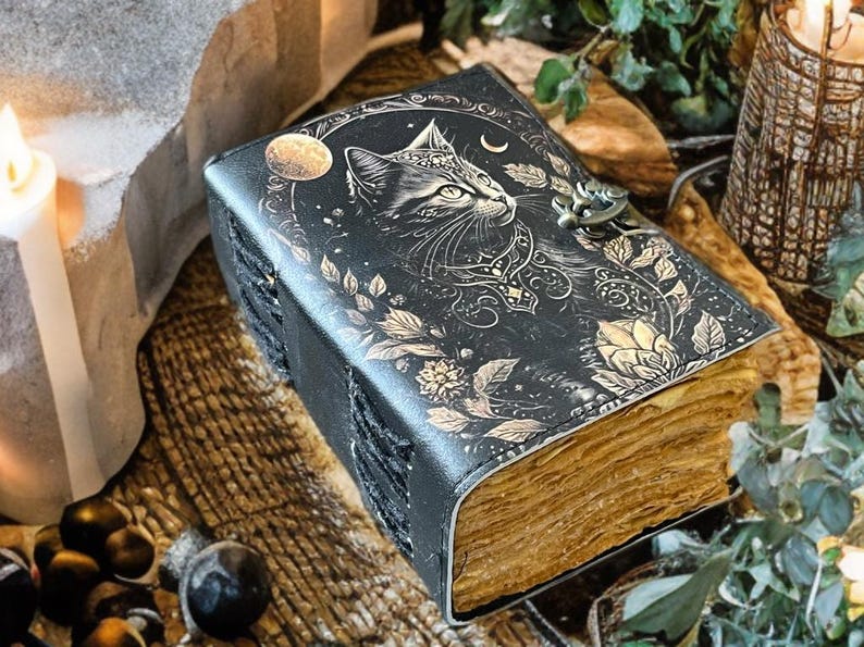 May include: A black leather journal with a cat design on the cover. The cover features an embossed cat with a crescent moon and floral accents. The journal has a metal clasp and aged-looking pages.