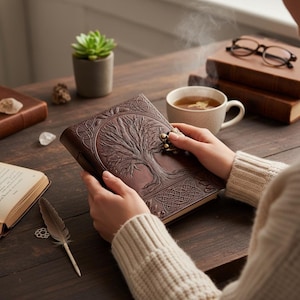 Könnte beinhalten: Ein braunes Lederjournal mit einem geprägten Lebensbaum-Design auf dem Einband. Das Journal hat einen Metallverschluss und wird von einer Person geöffnet gehalten. Eine Tasse Tee, Bücher und eine Sukkulente befinden sich im Hintergrund.