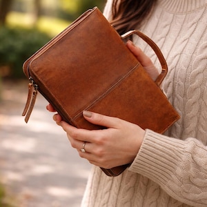 May include: A brown leather portfolio with a handle and zipper closure. The portfolio is held by a person wearing a cream-colored cable knit sweater. The leather has a natural grain and a stitched seam detail.
