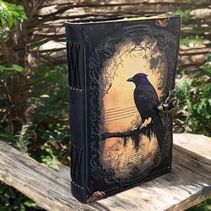 May include: A black leather journal featuring a raven illustration on the cover. The raven sits on a branch against a sepia background. The journal has a metal clasp and decorative stitching along the spine.