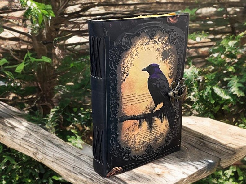 May include: A black leather journal featuring a raven illustration on the cover. The raven sits on a branch against a sepia background. The journal has a metal clasp and decorative stitching along the spine.