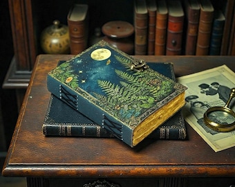 May include: A decorative journal with a celestial design featuring a full moon, stars, and fern leaves. The journal has a dark blue cover with gold-gilded edges and a metal clasp. The journal is stacked on top of another dark blue journal.