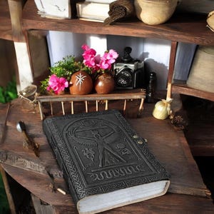 May include: A dark gray leather journal with an embossed design featuring a human figure, symbols, and a serpent. The journal is closed with a metal clasp. The background includes a wooden shelf with decorative items and pink flowers.