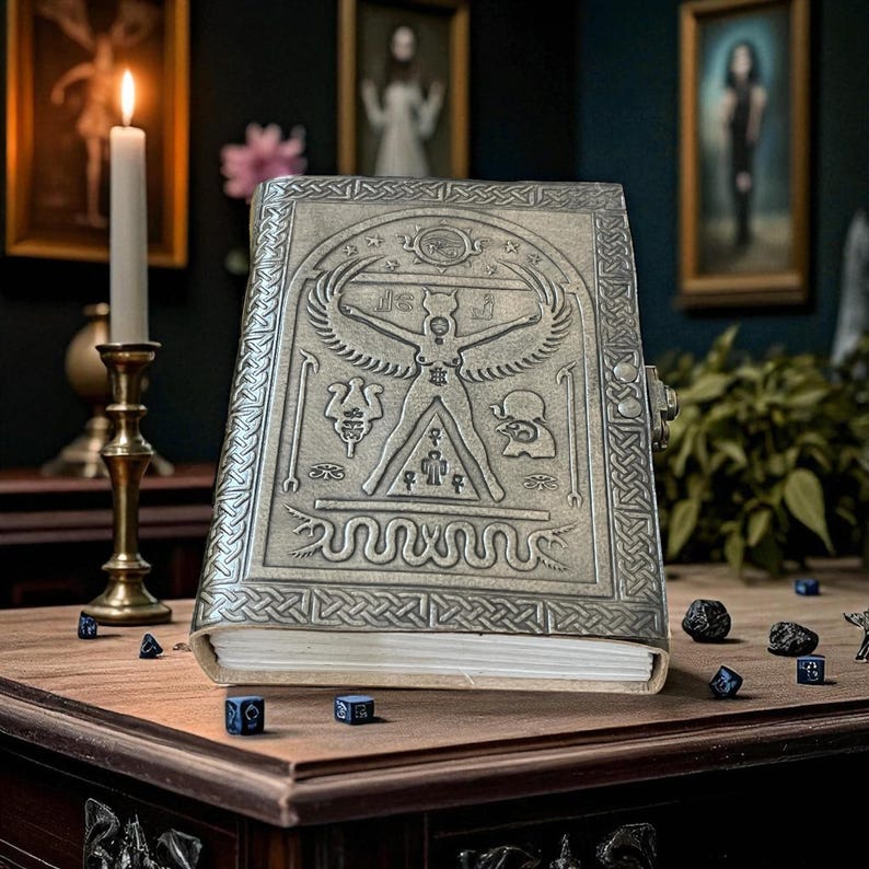 May include: A silver-colored, embossed journal with intricate designs, including a central figure with outstretched arms and wings. The journal is on a wooden table with dice scattered around. A lit candle and framed artwork are in the background.