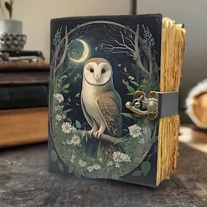 以下が含まれることがあります： フクロウのイラストが描かれた革張りのジャーナル。表紙には、白い花と三日月のある月明かりの森のシーンにいるメンフクロウの詳細な画像が描かれています。ジャーナルには金属製の留め金と古くなった紙の端があります。