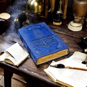 May include: A blue leather journal with embossed artwork featuring a hammer and wolf designs. The journal has a clasp closure and aged-looking pages. The cover also has text. The journal is on a wooden surface with other items.