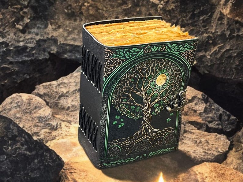 Puede incluir: Un diario de cuero negro con un dise&ntilde;o de &aacute;rbol de la vida en verde y dorado. El diario tiene un cierre de broche y un lomo decorativo. Las p&aacute;ginas est&aacute;n envejecidas con un tono dorado. El diario est&aacute; sobre una roca.