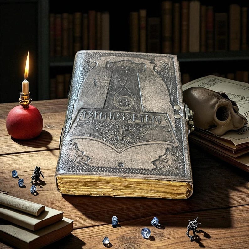 May include: A large, gray leather-bound book with an embossed design featuring a hammer and runes. The book is open, with gold-edged pages. A lit candle in a red holder, a skull, and small figurines are on the wooden table.