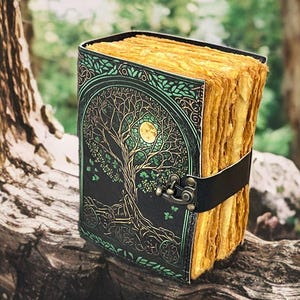 Puede incluir: Un diario encuadernado en cuero con un dise&ntilde;o de &aacute;rbol de la vida en la portada. La cubierta es negra con detalles verdes y dorados. Las p&aacute;ginas son envejecidas y el diario se asegura con una correa de cuero negro.