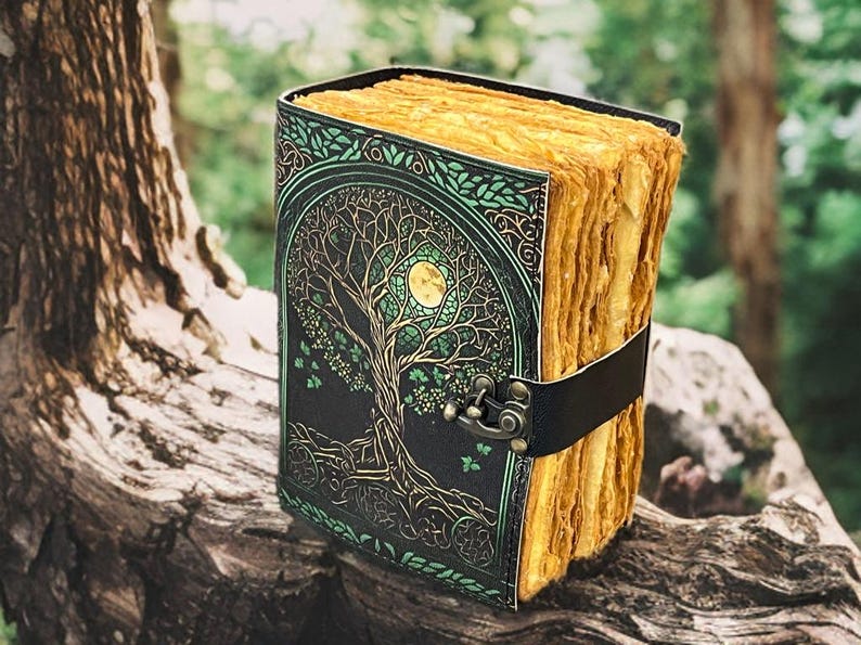 Puede incluir: Un diario encuadernado en cuero con un dise&ntilde;o de &aacute;rbol de la vida en la portada. La cubierta es negra con detalles verdes y dorados. Las p&aacute;ginas son envejecidas y el diario se asegura con una correa de cuero negro.