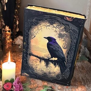 May include: A decorative book featuring a raven illustration. The book has a black cover with intricate detailing and a gold-coloured edge. The raven is perched on a branch against a forest backdrop.