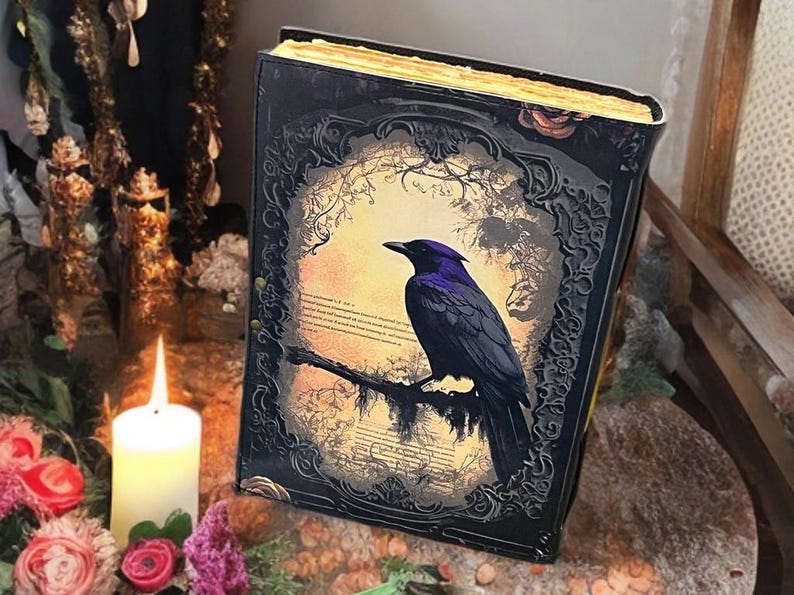 May include: A decorative book featuring a raven illustration. The book has a black cover with intricate detailing and a gold-coloured edge. The raven is perched on a branch against a forest backdrop.