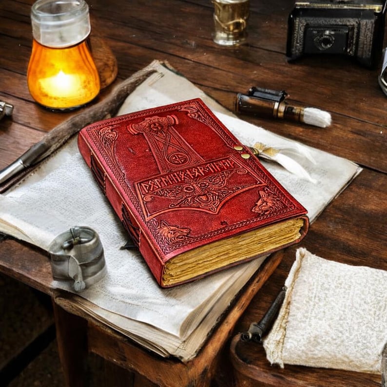 May include: A red leather-bound journal with embossed Norse designs, including a hammer and wolf heads. The journal is closed with a brass clasp and rests on a vintage paper. The pages have a yellowed, aged appearance. The journal is on a wooden table.