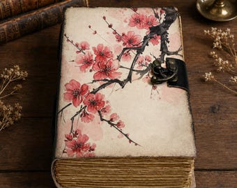 May include: A rectangular journal with a cream-colored cover featuring a black ink branch illustration with pink cherry blossoms. The journal has a dark, distressed edge finish, a black leather-like strap, and a metallic bronze-toned latch closure.